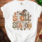 White t-shirt with "Spell Season" text, pumpkin, witch hat, and autumn leaves graphic.