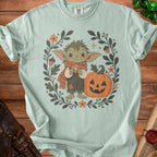 Green creature in scarf holding dice, with pumpkin and floral wreath.