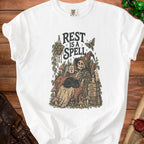 Skeleton reading, drinking coffee, with cat and text "REST IS A SPELL".