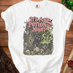 White t-shirt graphic with a black cat, potion, and spooky castle; text reads "It's a Potion Night."