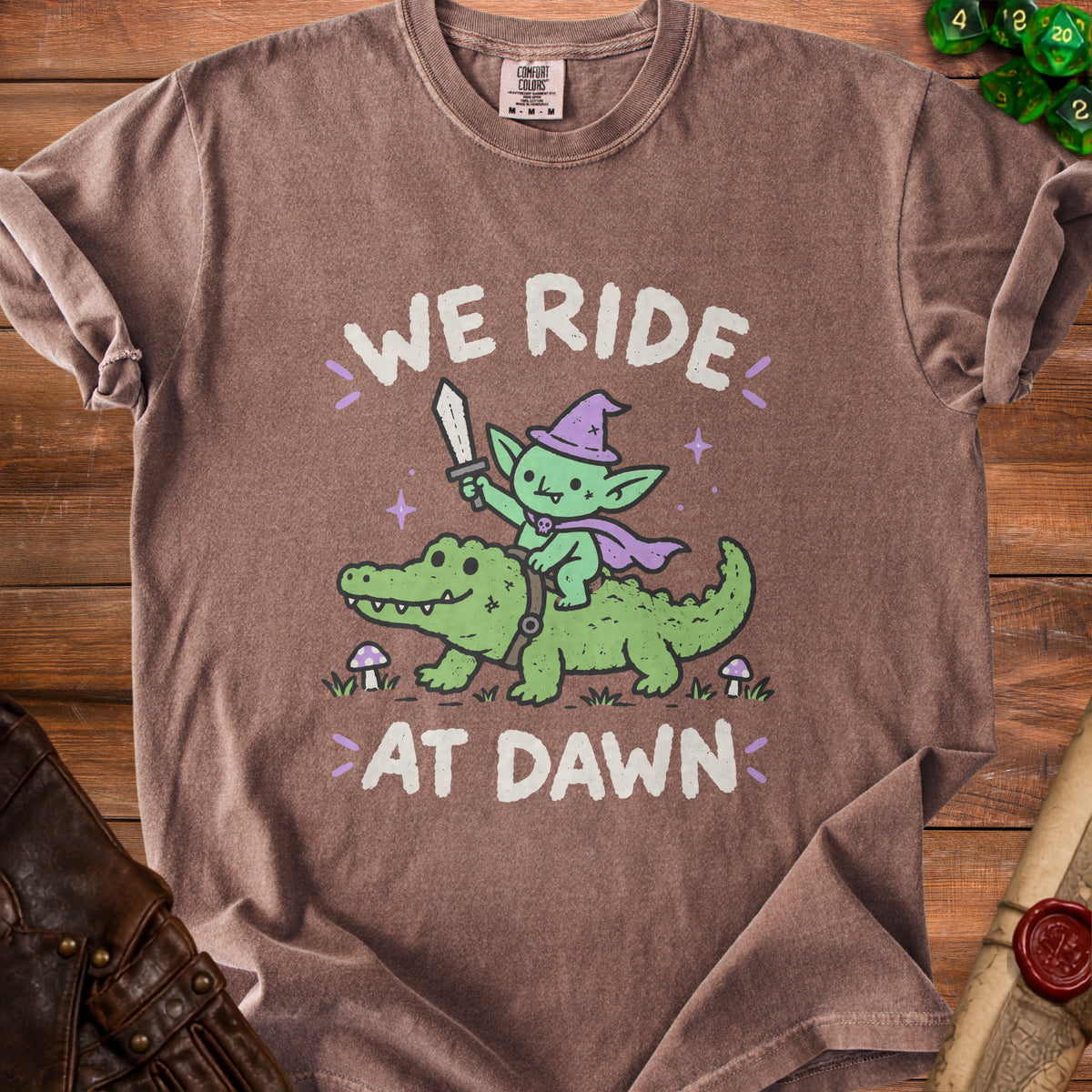 Goblin riding alligator with sword, text "WE RIDE AT DAWN" on brown shirt.