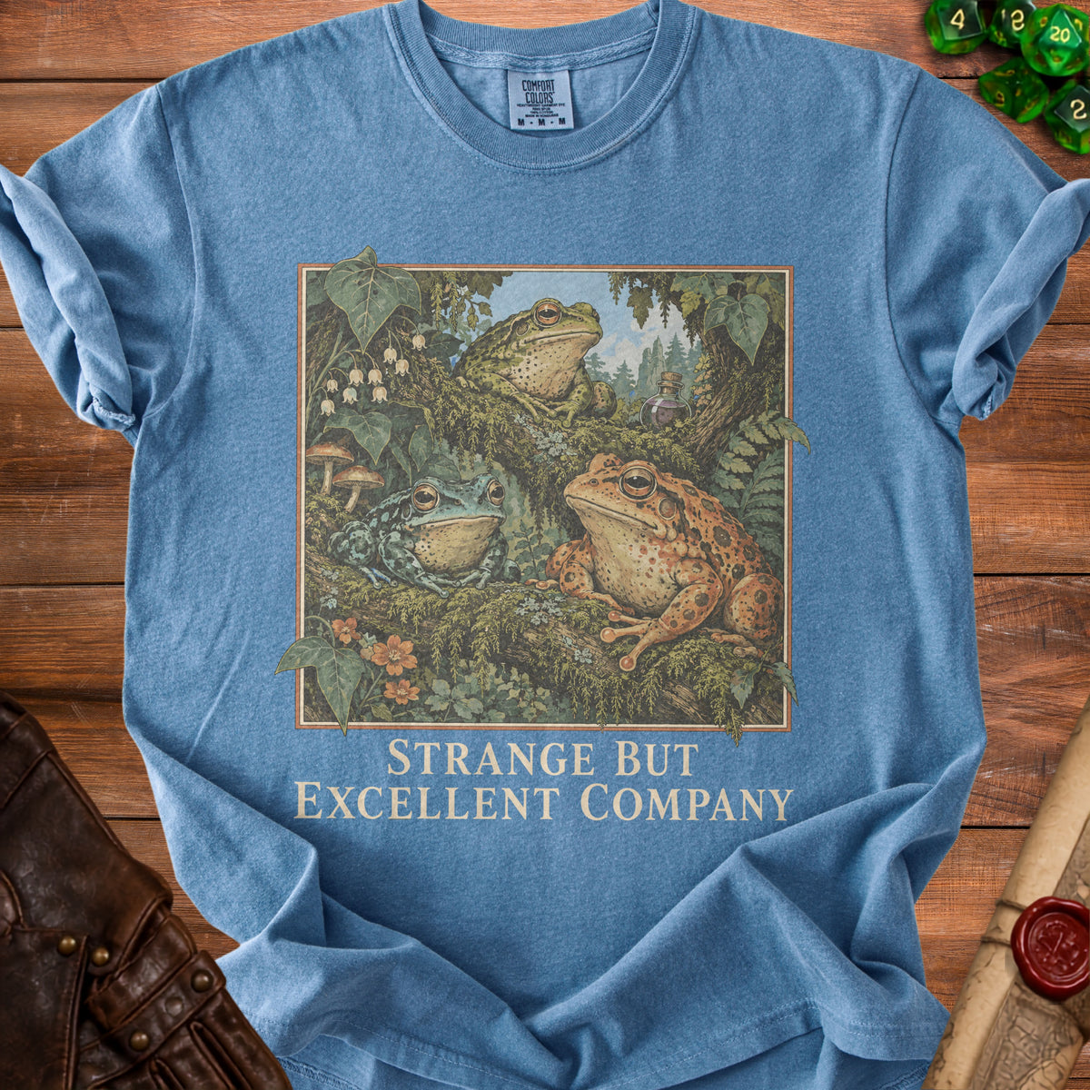 Blue t-shirt with an illustration of three frogs in a forest setting and the text "Strange But Excellent Company".