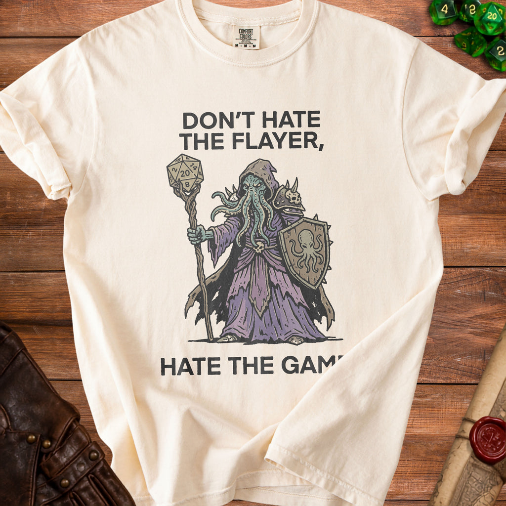 Cream t-shirt with a creature holding a d20 and shield, text reads "Don't hate the flayer, hate the game."