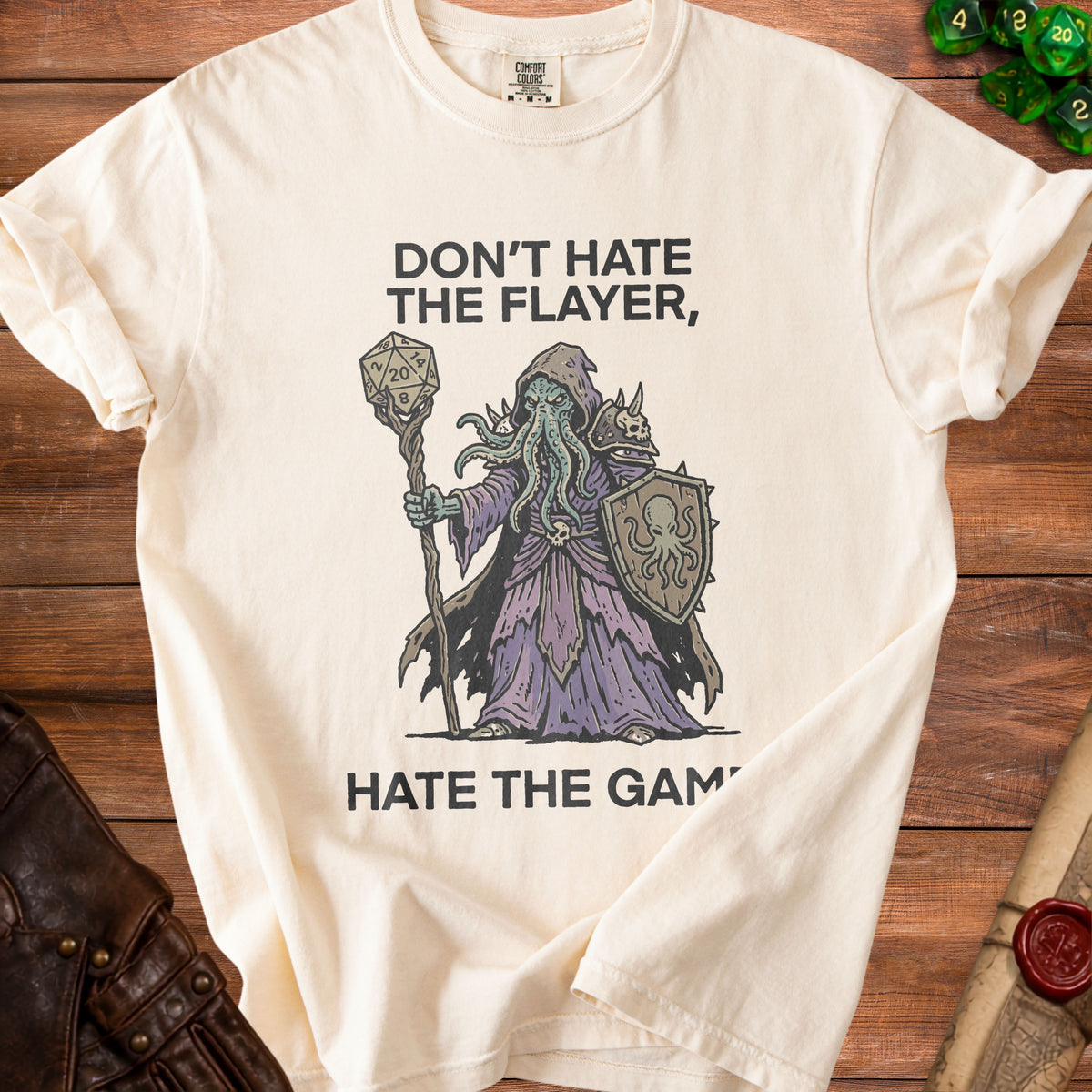 Cream t-shirt with a creature holding a d20 and shield, text reads "Don't hate the flayer, hate the game."