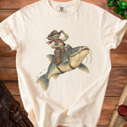 Goblin cowboy rides a catfish on a cream t-shirt with a weathered wood background.