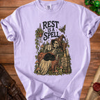 Skeleton in armchair with cat, holding "STILL HERE" mug, text "REST IS A SPELL".