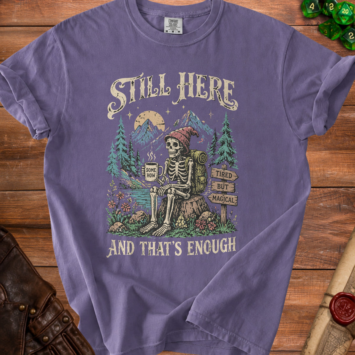 Purple t-shirt with skeleton camping graphic and text: "Still Here, Some How, Tired But Magical, And That's Enough."