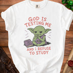 White t-shirt with text "GOD IS TESTING ME AND I REFUSE TO STUDY" and a goblin.