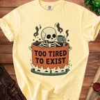 Yellow t-shirt with skeleton in cauldron graphic and text "TOO TIRED TO EXIST".
