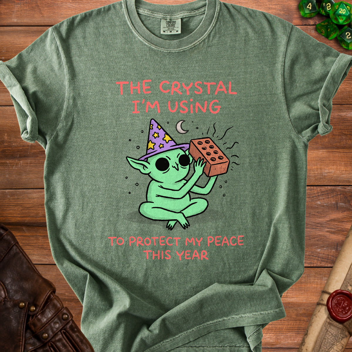 Green t-shirt with a cartoon goblin in a wizard hat holding a brick. Text reads "The crystal I'm using to protect my peace this year."