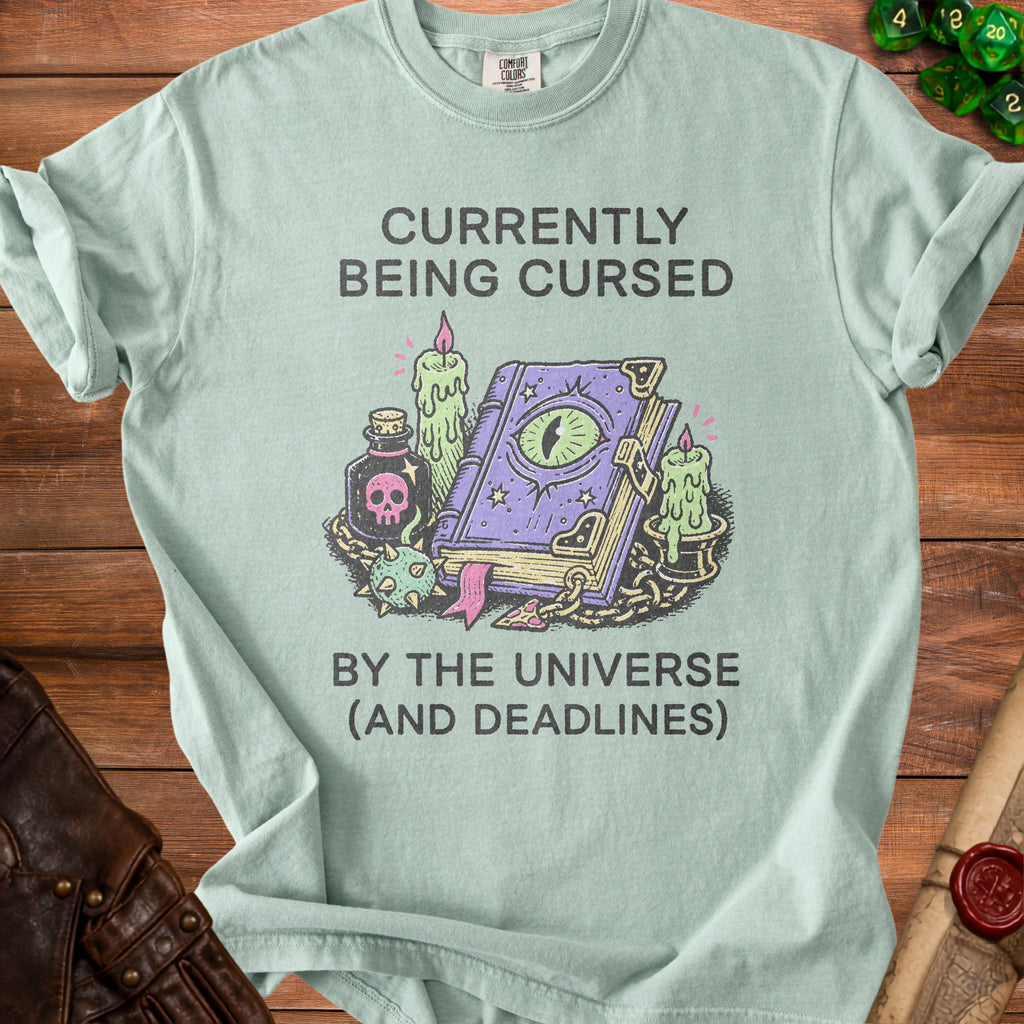 Light blue t-shirt with "Currently Being Cursed By The Universe (And Deadlines)" text and magical illustration.