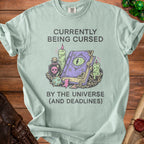 Light blue t-shirt with "Currently Being Cursed By The Universe (And Deadlines)" text and magical illustration.