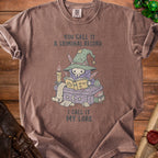 Brown t-shirt with graphic of a skeleton witch reading a book titled "I Call It My Lore."
