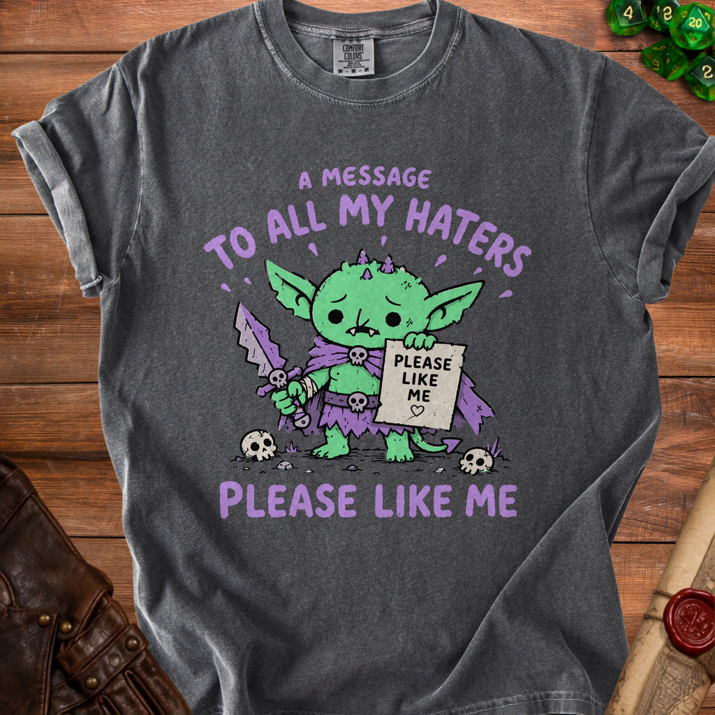 Dark gray t-shirt featuring a sad green goblin with a sword holding a "Please Like Me" sign.