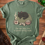 Green t-shirt with illustration of sleeping cat, coffee, popcorn, and game controller. Text reads "I'm not lazy, I'm in power-saving mode."