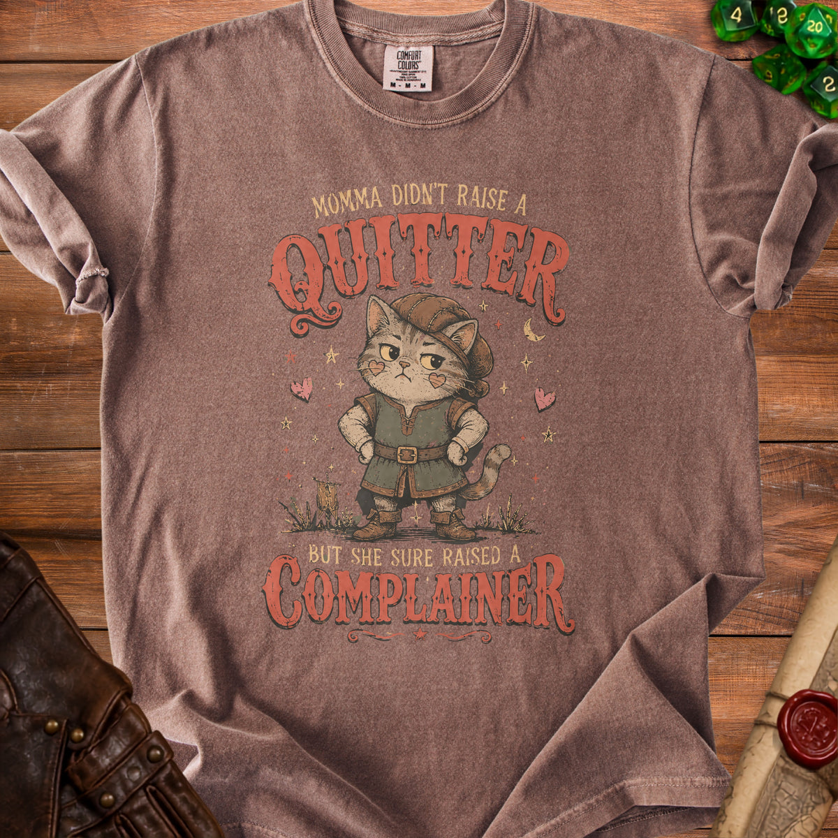 Brown t-shirt with a cartoon cat wearing a hat and tunic, stating "Momma didn't raise a quitter but she sure raised a complainer."