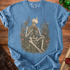 Skeleton holding a flower, surrounded by plants, on a blue t-shirt.