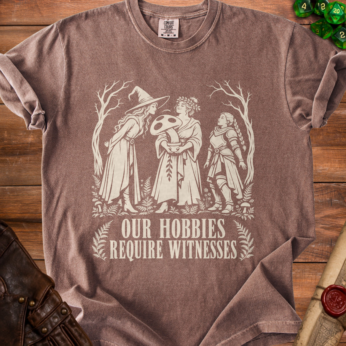 Brown t-shirt with cream graphic of three women holding a mushroom, text reads "OUR HOBBIES REQUIRE WITNESSES".