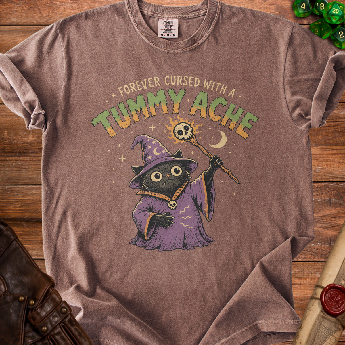 Brown t-shirt with a black cat wizard holding a skull staff, text "FOREVER CURSED WITH A TUMMY ACHE".