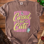 Brown t-shirt with "Cursed but Cute" text, a cat, and dice.