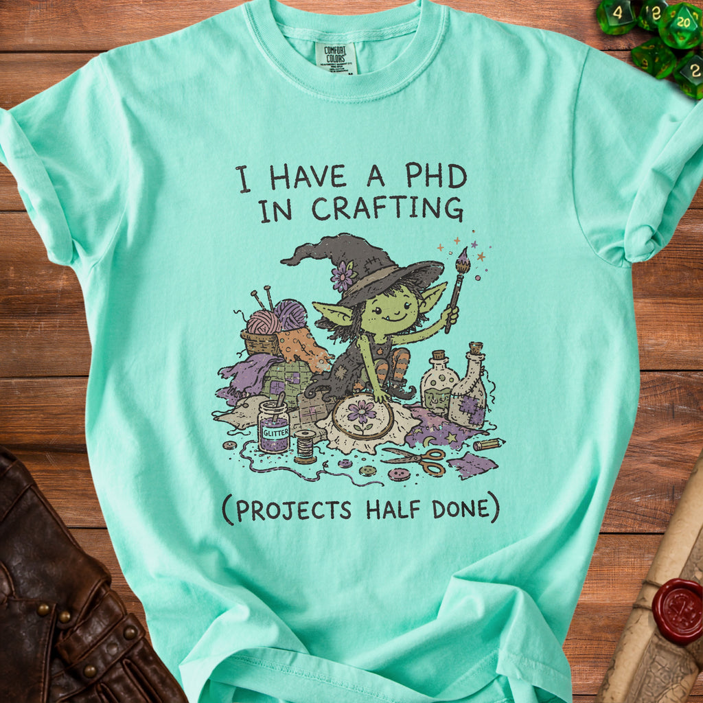 Teal t-shirt with a drawing of a goblin crafting surrounded by supplies and text.