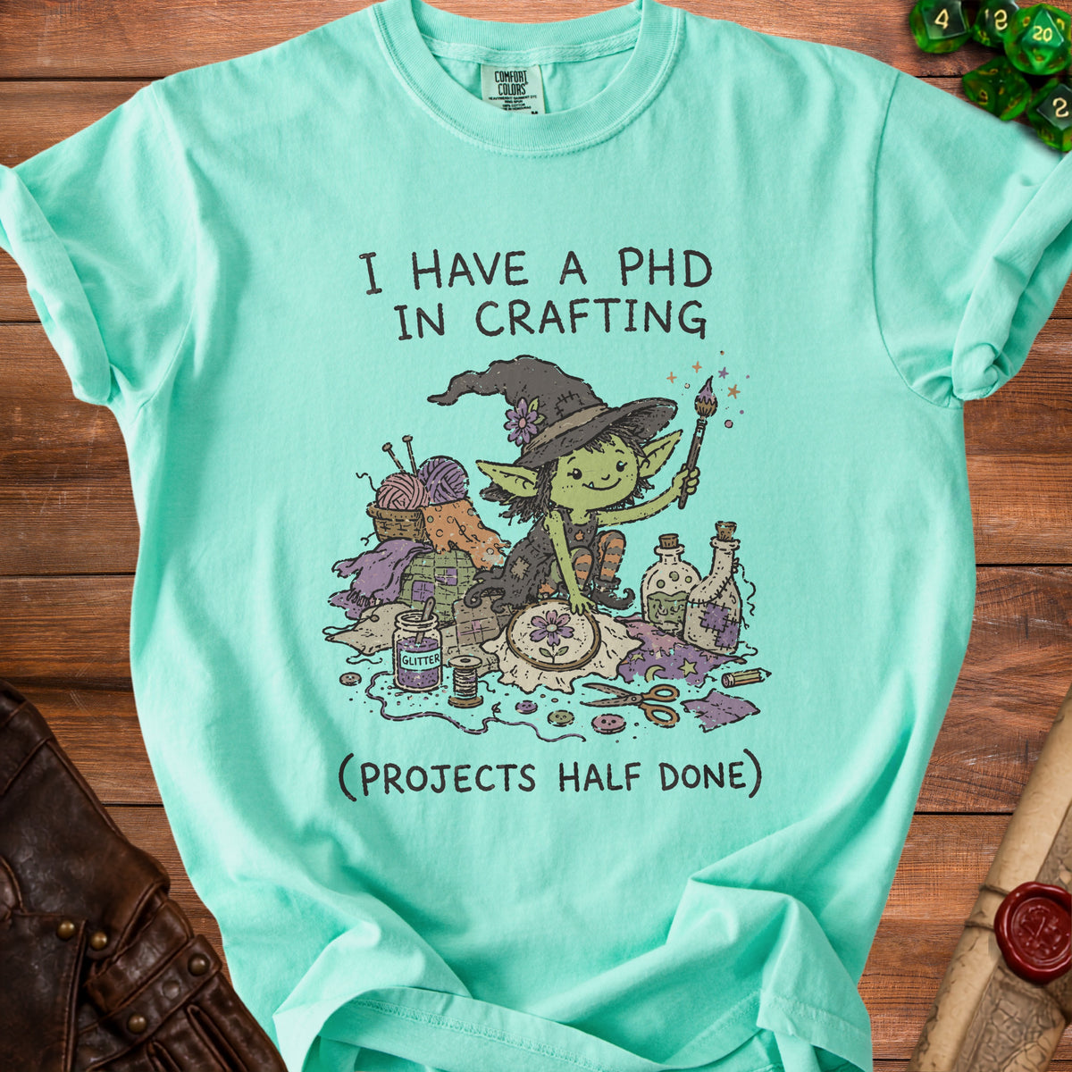 Teal t-shirt with a drawing of a goblin crafting surrounded by supplies and text.