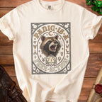 Cream t-shirt with a graphic of a raccoon, a d20 die, and the text "Panic is a Free Action".