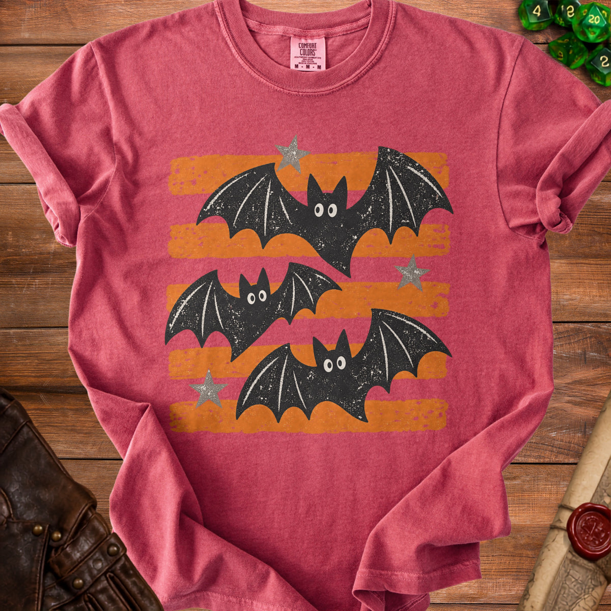 Pink t-shirt with a graphic of three black bats with white eyes on orange and pink stripes.