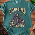 Skeleton wizard with text "DEAD TIRED STILL CASTING" on a teal t-shirt.