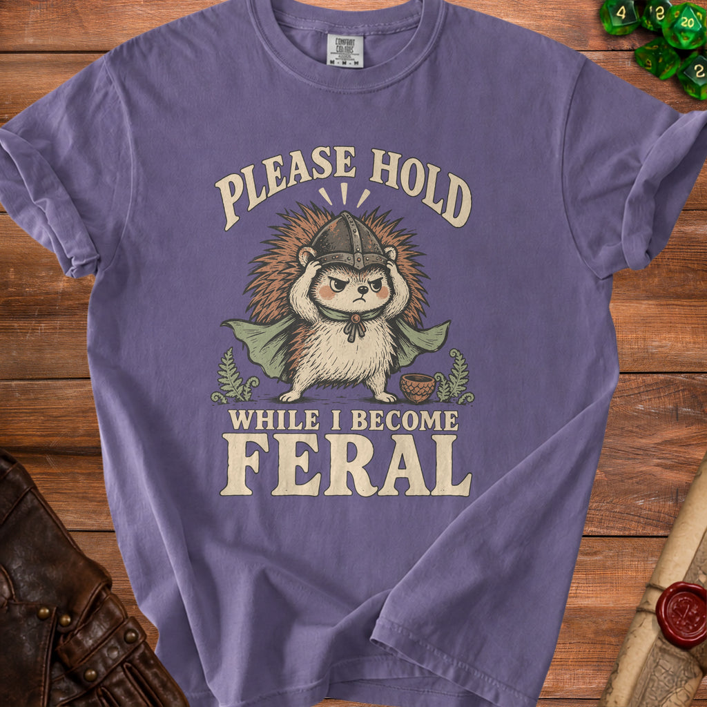 Purple t-shirt with a hedgehog wearing a helmet and cape, and text "PLEASE HOLD WHILE I BECOME FERAL".