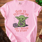 Pink t-shirt with graphic of grumpy goblin reading "How to Scheme" book and text "GOD IS TESTING ME AND I REFUSE TO STUDY."