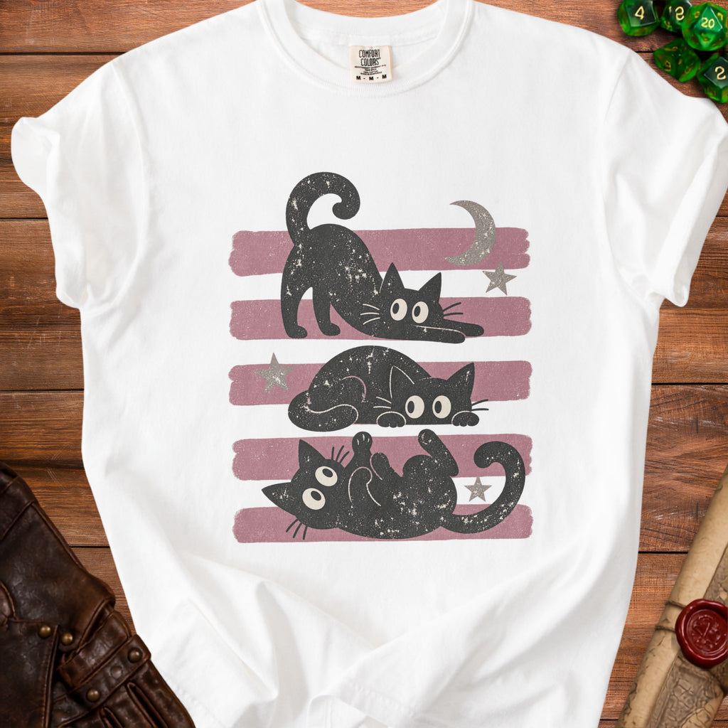 White t-shirt with three playful black cats on a striped background with moon and stars.