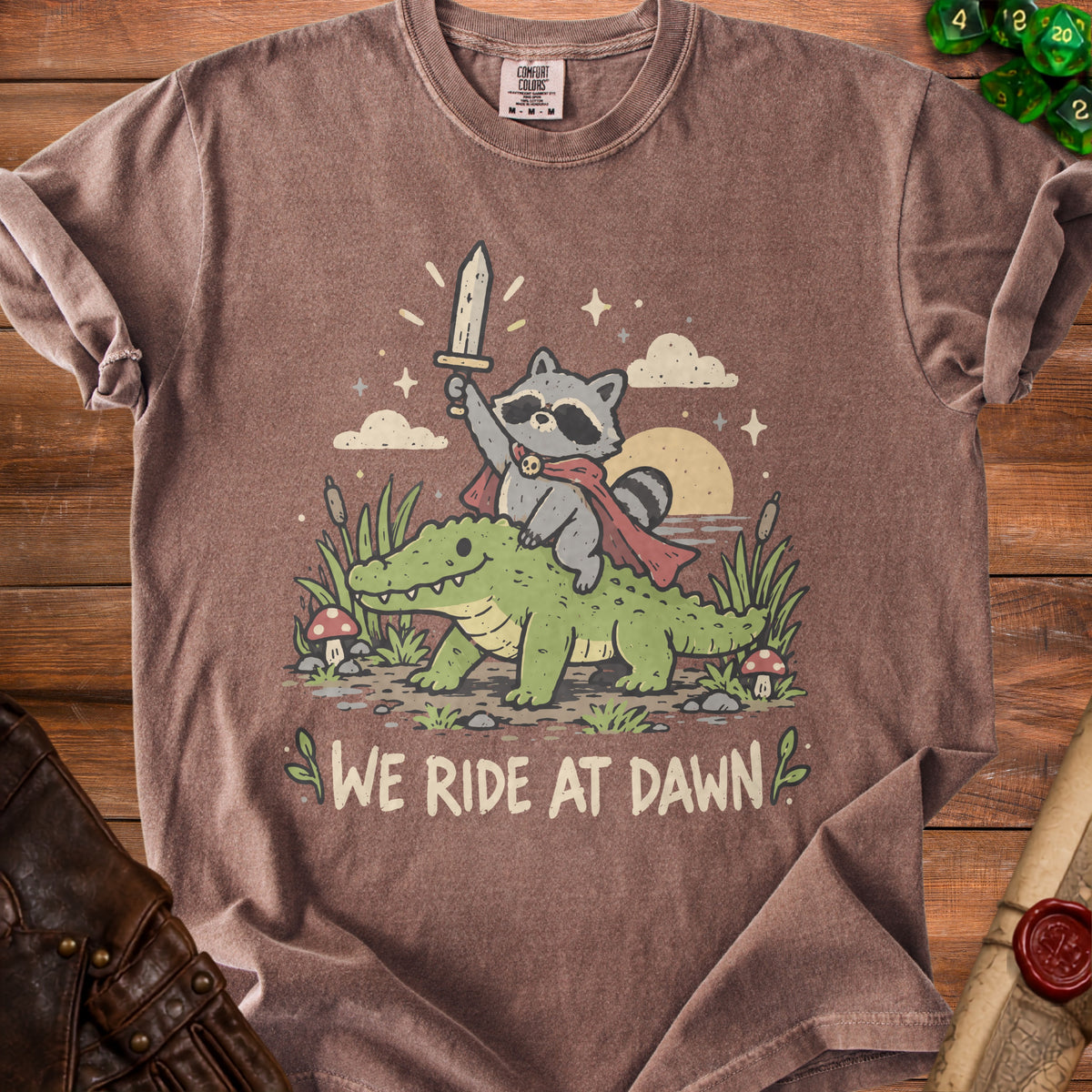 T-shirt with a raccoon riding an alligator and the text "WE RIDE AT DAWN".