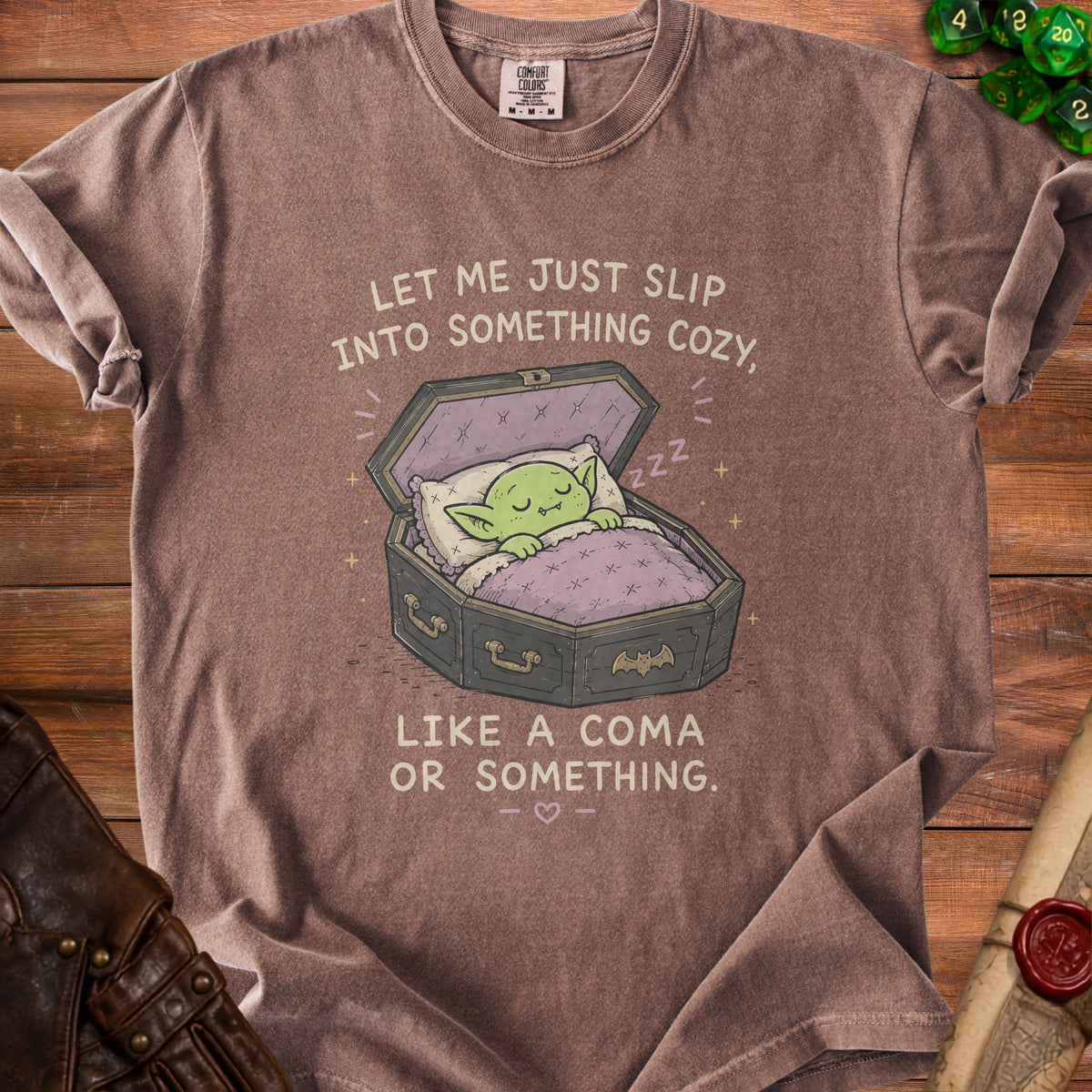 Brown t-shirt with a graphic of a green creature sleeping in a coffin. Text reads: "Let me just slip into something cozy, like a coma or something."