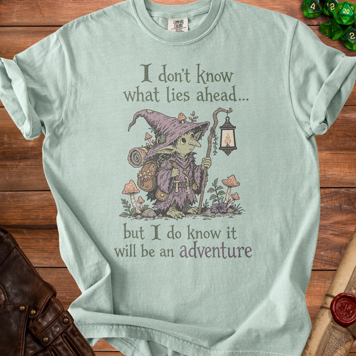 Light teal t-shirt with illustration of a goblin holding a lantern and the text "I don't know what lies ahead... but I do know it will be an adventure."