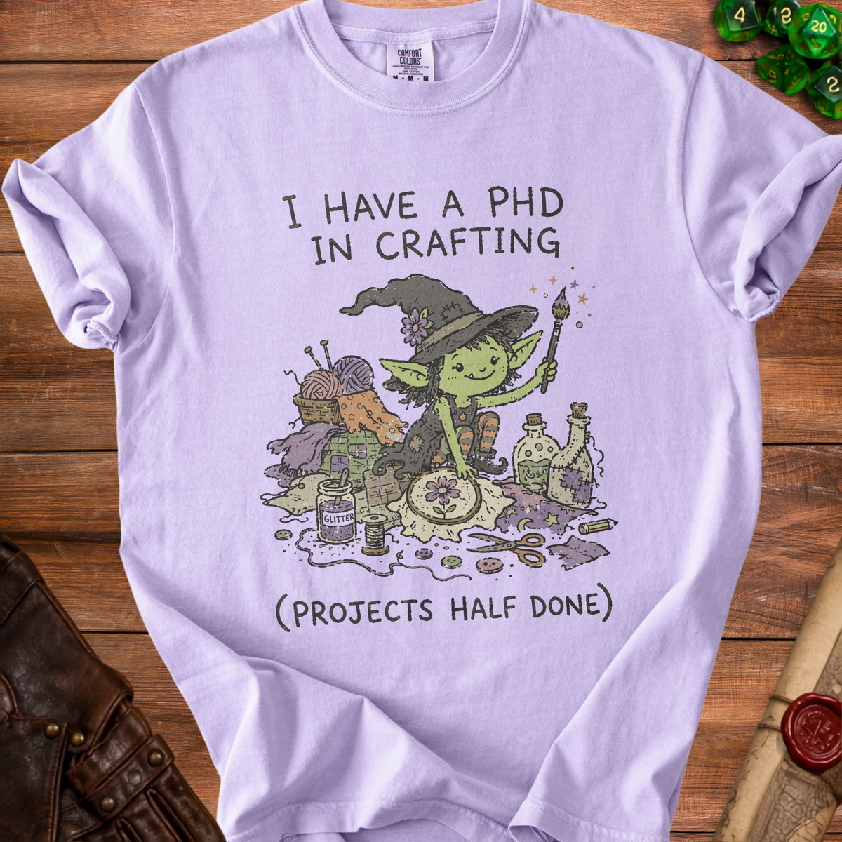 Lavender t-shirt with a graphic of a goblin with crafting supplies and text.