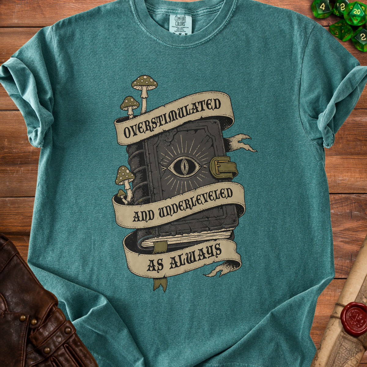 T-shirt graphic of a spellbook with an eye, mushrooms, and text "OVERSTIMULATED AND UNDERLEVELED AS ALWAYS".