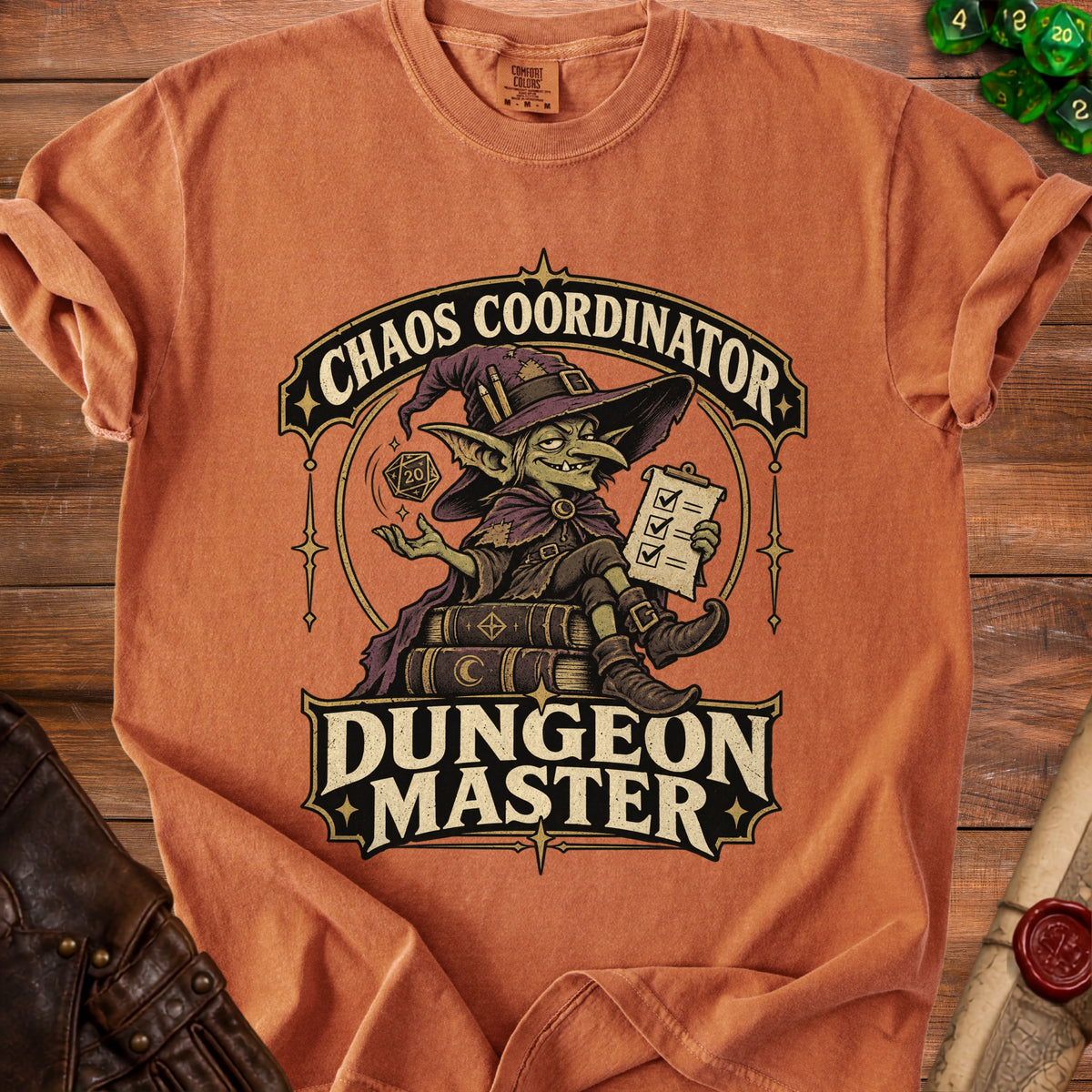 An orange t-shirt displays a graphic of a goblin wizard sitting on spellbooks, holding a clipboard and dice, with the text Chaos Coordinator Dungeon Master.