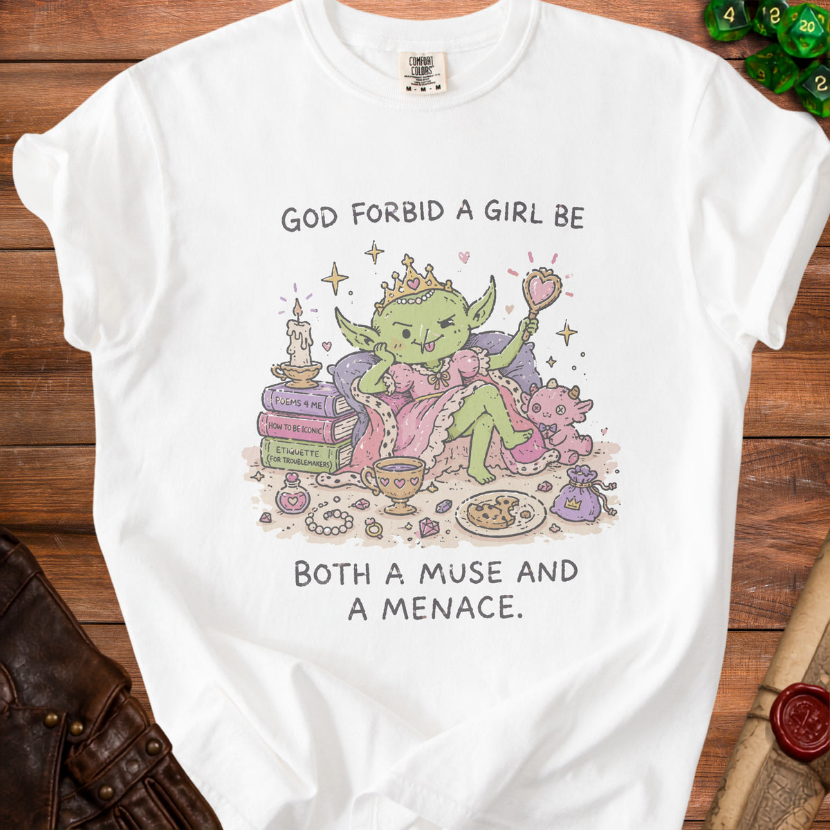 White t-shirt with a graphic of a crowned goblin girl saying "God forbid a girl be both a muse and a menace."