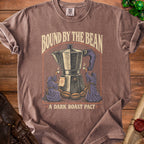 Brown t-shirt graphic with moka pot, robed figures, and text "Bound by the Bean, A Dark Roast Pact."
