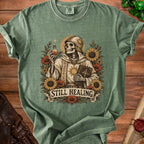 Skeleton in religious attire holding potion and book, with flowers and banner reading "STILL HEALING".