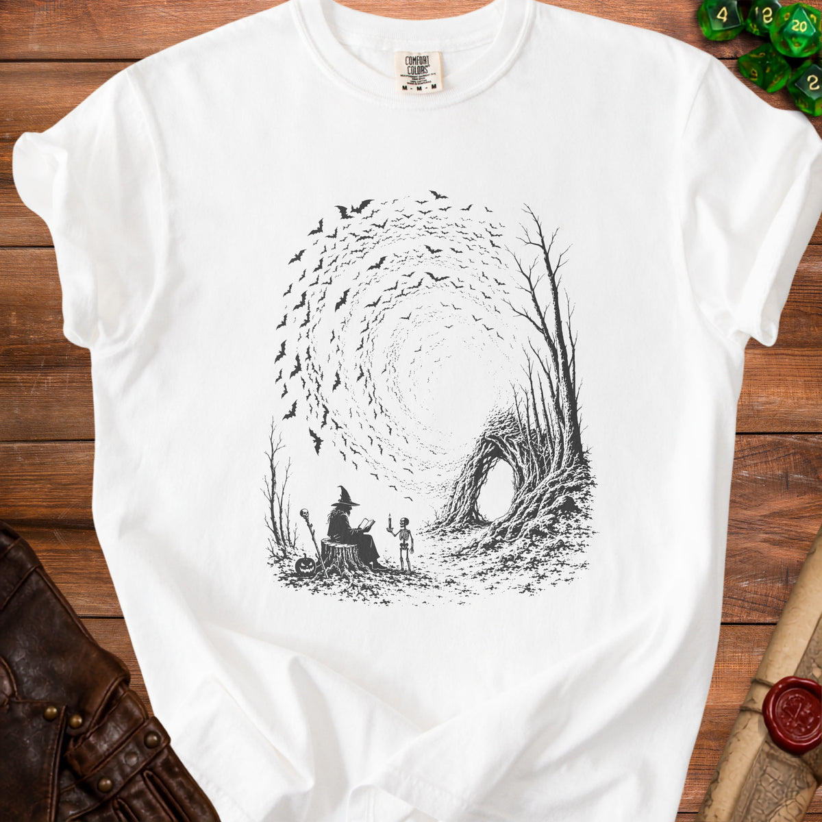 White t-shirt with a witch reading to a skeleton, surrounded by bats and trees.
