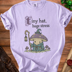 Lavender t-shirt with cartoon mushroom wearing a hat, holding a lantern, and text "Tiny hat, huge stress".