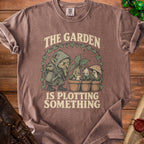 Brown t-shirt with graphic of a goblin tending plants and text "The Garden Is Plotting Something".