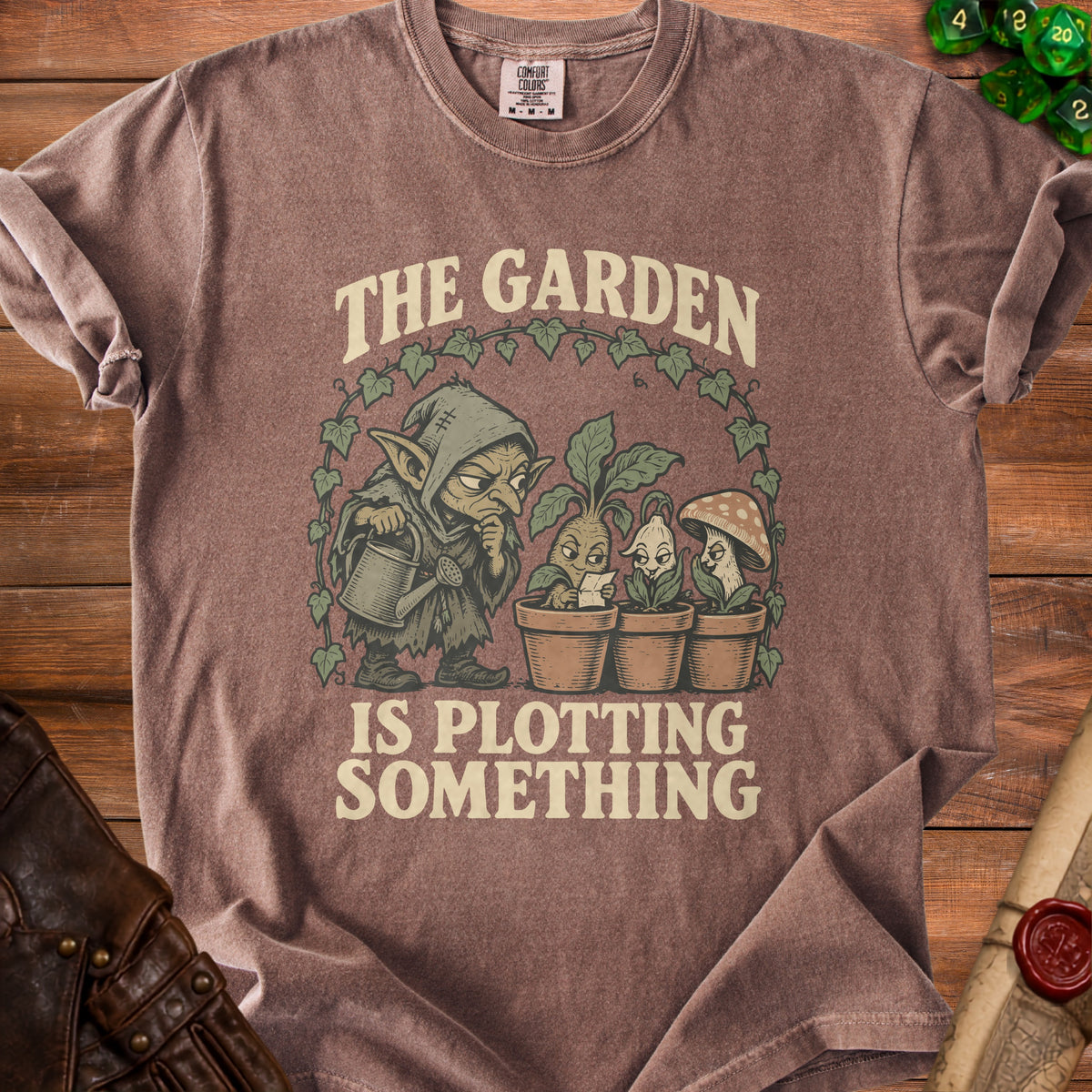 Brown t-shirt with graphic of a goblin tending plants and text "The Garden Is Plotting Something".