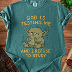 Teal t-shirt with cartoon creature and text "God is testing me and I refuse to study".