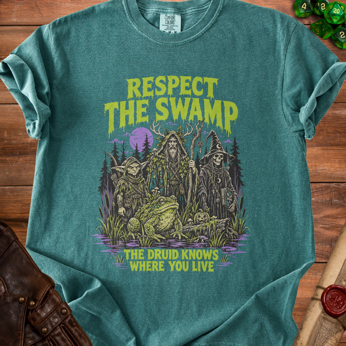 Green t-shirt graphic of a swamp with a toad, a druid, a goblin, and a skeleton wizard. Text reads "RESPECT THE SWAMP THE DRUID KNOWS WHERE YOU LIVE".
