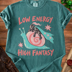 A teal t-shirt featuring a graphic of a knight sleeping on a snail shell, with pink text reading "LOW ENERGY HIGH FANTASY".