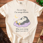 Cream t-shirt with cartoon penguin on cloud, text "I'm not lazy, I'm energy-efficient. Why waste energy when I can save it?".