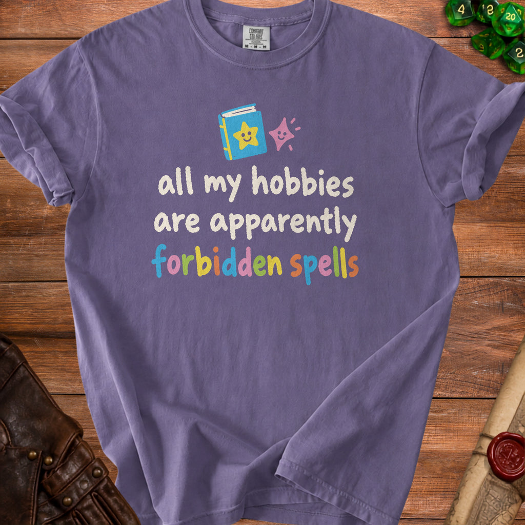 Purple t-shirt with text "all my hobbies are apparently forbidden spells" and cartoon book graphic.